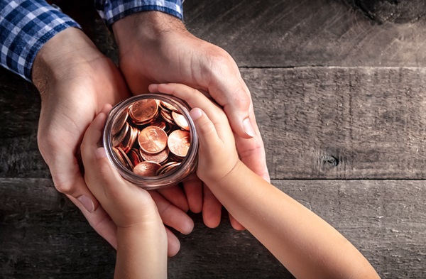 Adult and child hands holding a glass jar filled with coins over a wooden table.